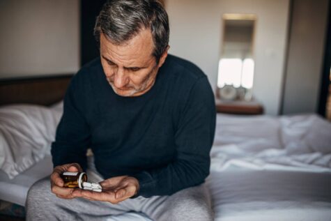 Älterer Mann mit Tabletten, am Bett sitzend.